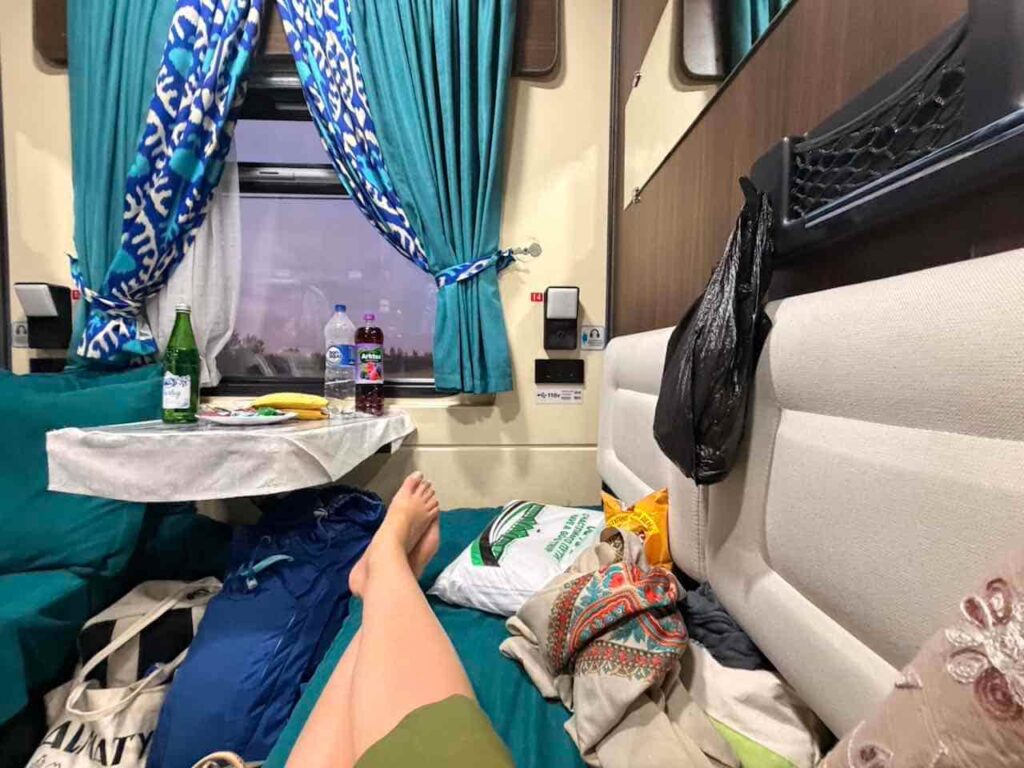 Auf dem Bett im Nachtzug liegend. Blick auf den Tisch mit Getränken und Lebensmitteln und aus dem Fenster.